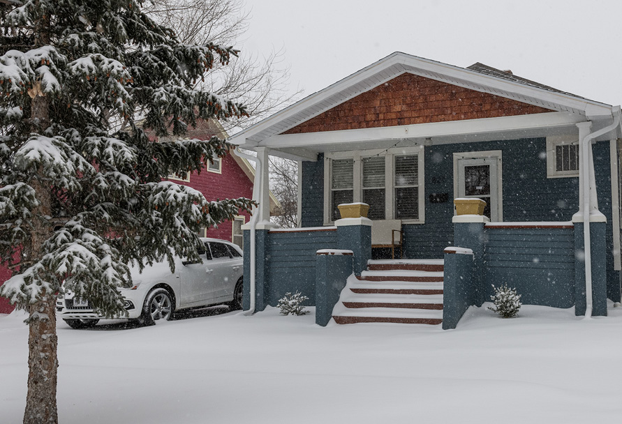 Snowy view of a blue house with a white car parked next to it and a pine tree covered in snow in the foreground
